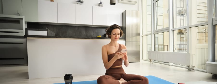 Yoga and sport at home concept. Young woman sitting in bright rome with mobile phone, doing workout on rubber mat, searching for exercises online on smartphone app, using sports equipment.