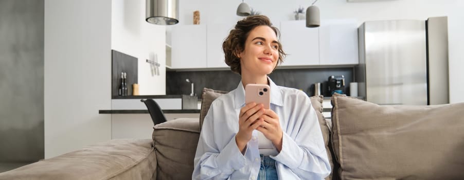 Image of brunette smiling woman with smartphone, sitting at home on sofa, looking aside with happy, thoughtful face, chatting on phone, using mobile app.