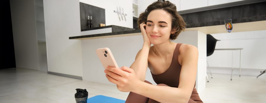Technology and sport concept. Fitness girl looking at her smartphone, sitting in workout clothes at home, watching exercise videos on mobile phone.