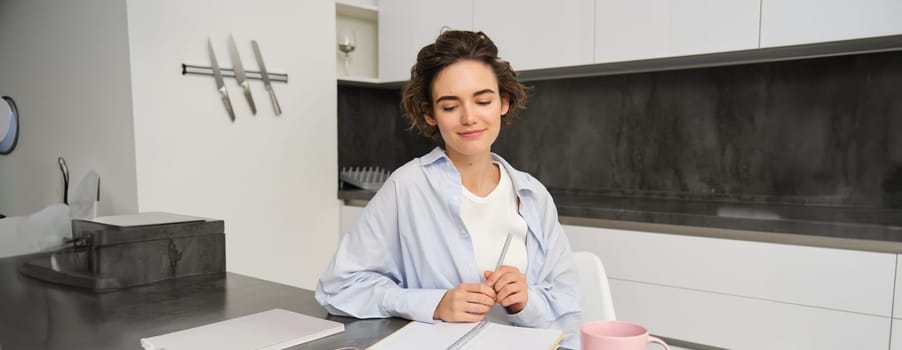 Portrait of young woman working from home, writing down information in notebook, taking notes, sitting in kitchen and studying, student doing homework.