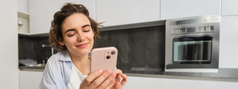 Portrait of modern brunette woman holding mobile phone and smiling. Girl sits in kitchen at home with smartphone, using app to order delivery, does online shopping.