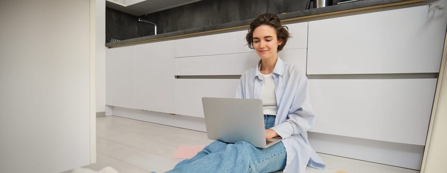 Portrait of girl freelancer, sits at home on floor and works with laptop, surrounded with piles of documents, smiles while looks at computer screen.