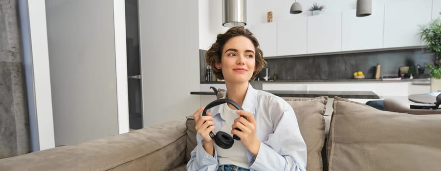 Portrait of beautiful modern girl on sofa, sits with headphones in hands and looks aside, listens music, enjoys e-book in earphones, spends time at home.