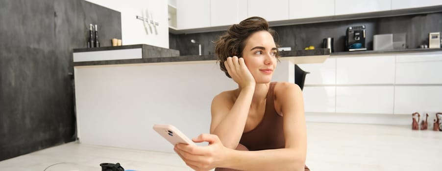 Portrait of sporty girl with mobile phone, sitting on yoga mat, using smartphone during workout at home, watching fitness turorial on telephone. Sport and wellbeing concept