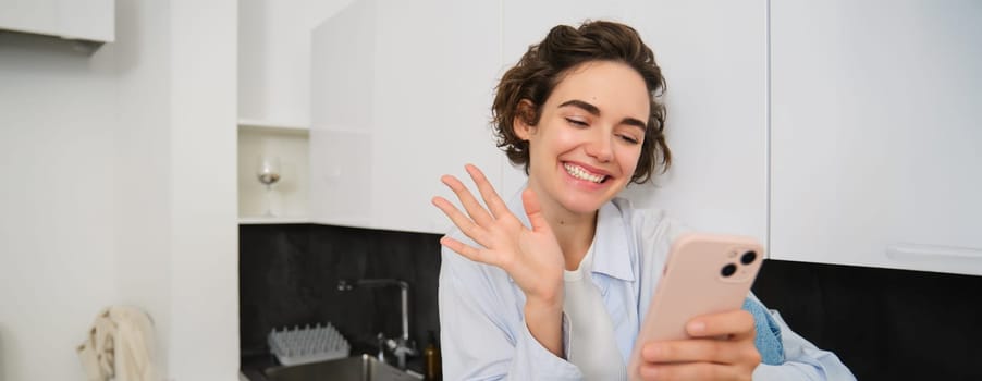 Portrait of friendly smiling brunette woman, sitting at home in kitchen, looking at her smartphone camera, video chats and waves at mobile phone, saying hello to someone.