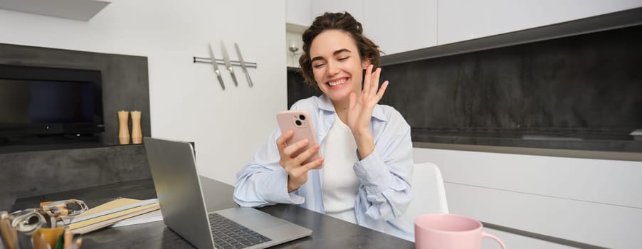 Happy young woman says hello, waves hand at smartphone and smiles, video chats with someone on mobile phone app.