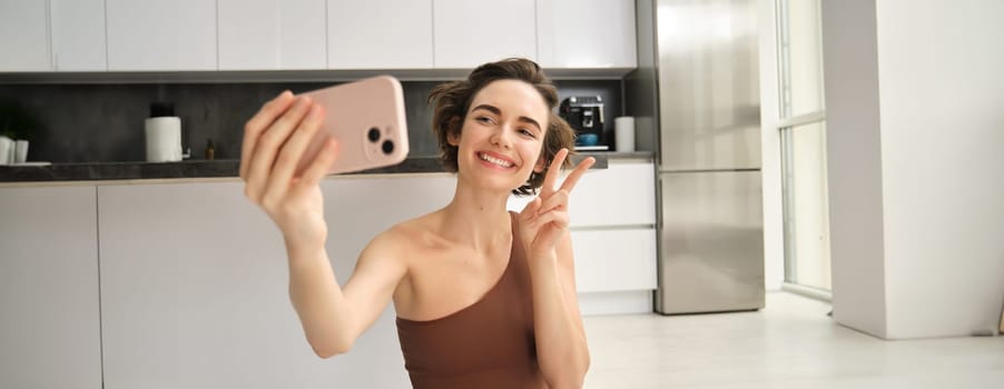 Smiling fitness girl, takes selfie during workout at home, posing for photo and holding smartphone, workout from home, recording vlog of her yoga training session.