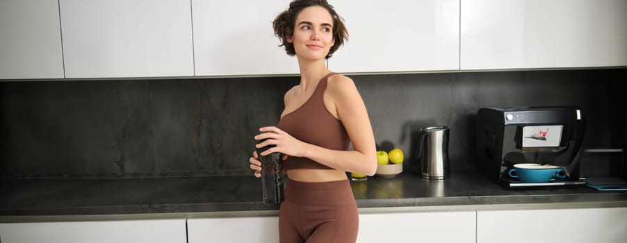 Portrait of smiling fitness girl, wearing sportswear for jogging, workout training, standing in kitchen and drinking water from sports bottle, looking fit and happy.