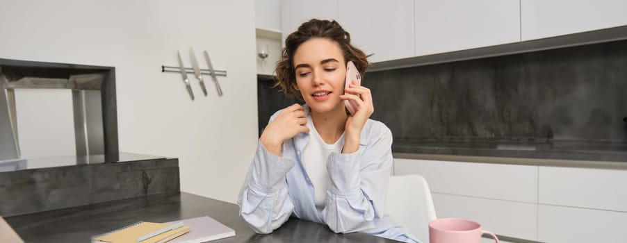 Portrait of young woman, 25 years old, holding smartphone, talking on mobile phone, sitting in kitchen at home.