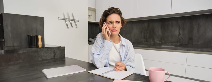 Portrait of woman working from home, calling someone on mobile phone, looking confused while holding pen and trying to write down something, sitting with puzzled face in kitchen.