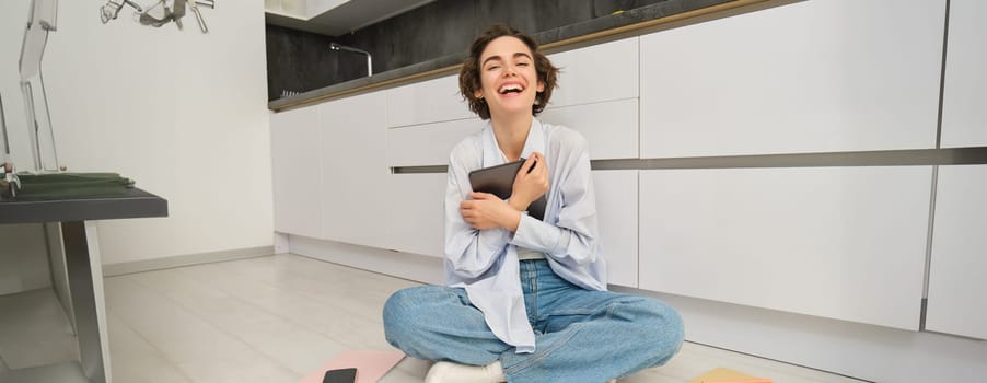 Portrait of happy woman with tablet, sitting on floor, laughing and smiling, working from home on remote, studying online.