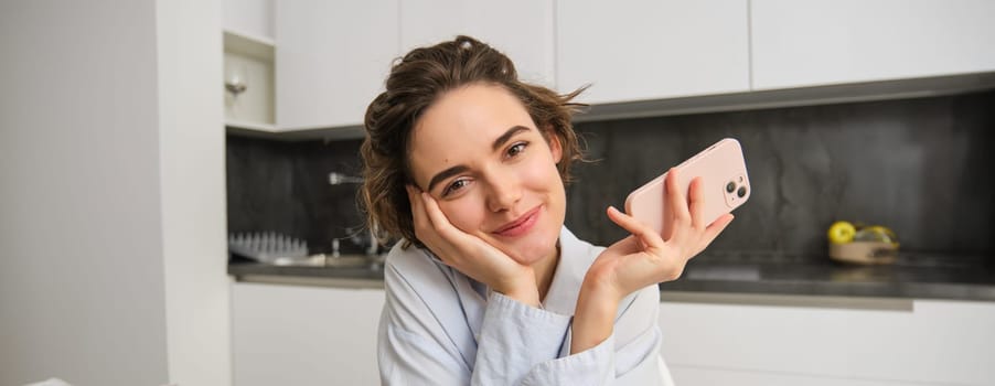 Candid smiling woman, holding smartphone in her kitchen, using mobile phone and looking happy.