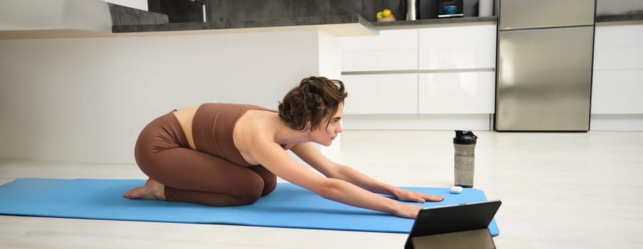 Image of young sportswoman does yoga at home, watches gym instructor tutorial video on tablet, stretches body, fitness training exercises, pilates class online from home.