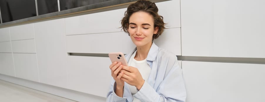 Indoor shot of happy girl with smartphone, sits on floor at home and uses mobile phone, watches video in app, chats with friends in applicatiion.