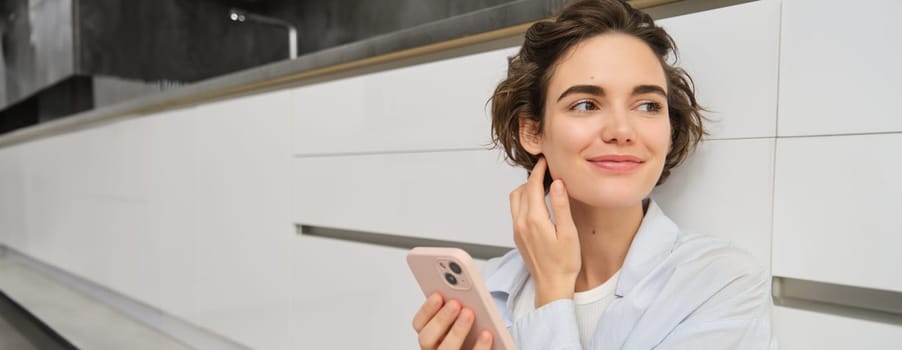 Close up portrait of happy woman with a phone at home, she touches her face and smiles lightly, uses mobile application. Technology and people concept.