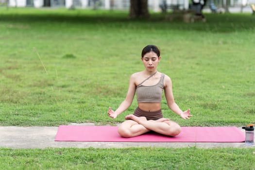 Asian woman in sporty outfit relaxing meditating feeling zen like on fitness mat in public park outdoor. Healthy active lifestyle.
