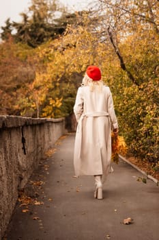 autumn woman in a red beret, a light coat and a red skirt, against the backdrop of an autumn park with yellow leaves in her hands.