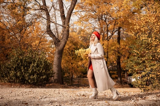 autumn woman in a red beret, a light coat and a red skirt, against the backdrop of an autumn park with yellow leaves in her hands.