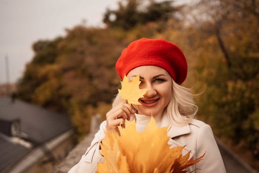 autumn woman in a red beret, a light coat and a red skirt, against the backdrop of an autumn park with yellow leaves in her hands.