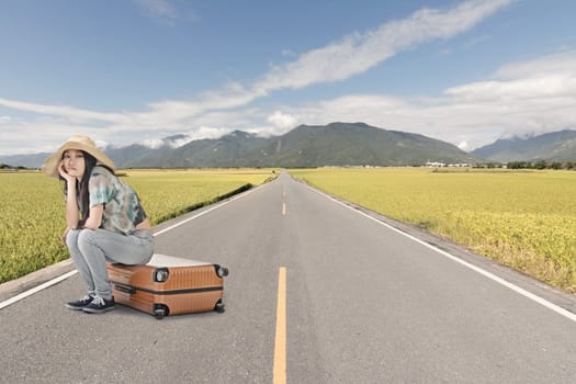 Asian woman thinking and sitting on a luggage on the ground of road in countryside.