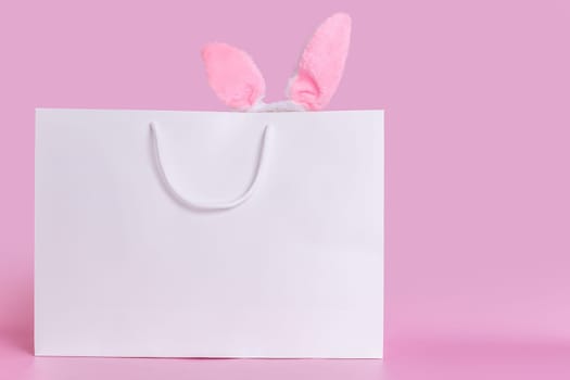 A big white paper bag on a pink background with pink fluffy ears , standing on a pink background. Copy space. Happy Easter concept