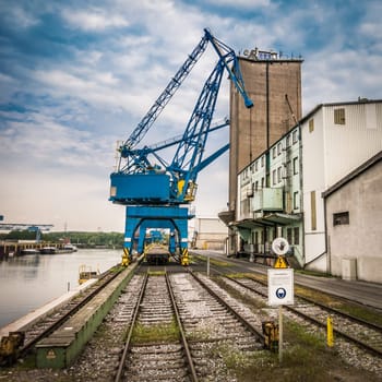 A blue harbour crane in an inland port in Germany,a few warehouses,rails and a blue sky with a few clouds.