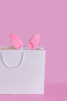 The ears of the Easter bunny in a large white paper gift bag standing on a pink background. Space for text. Vertical