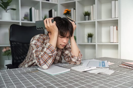 A frustrated young man sits at a desk, overwhelmed by financial management tasks with a calculator, phone, and scattered documents.