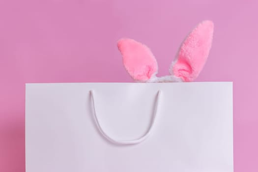 Close-up of a white paper bag on a pink background, with fluffy pink Easter bunny ears sticking out. Copy space. Happy Easter concept