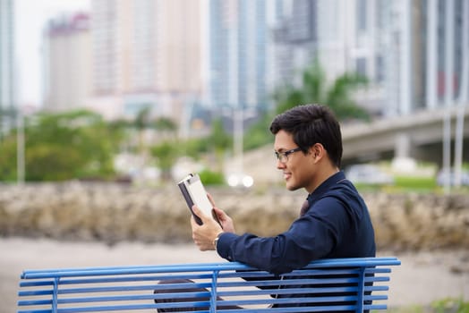 Portrait of chinese businessman sitting on bench in Panama city with skyline in background, reading emails on tablet computer