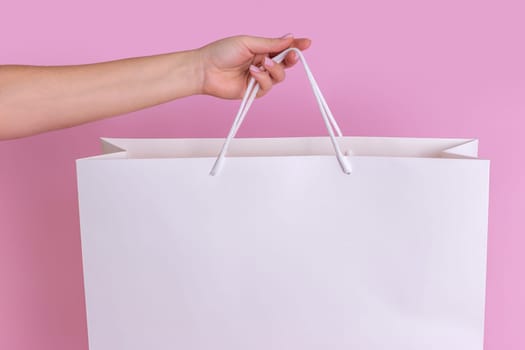 A close-up of a womans hand holding a large white transparent gift bag made of kraft paper for shopping after shopping, highlighted on a pastel pink background. layout. Advertising layout of the delivery concept