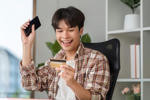 A joyful young man celebrates successful financial management using a smartphone and credit card, showcasing happiness.