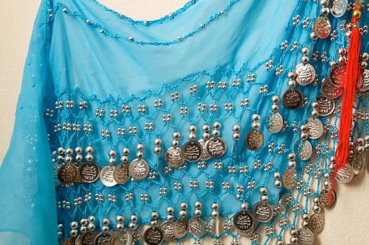 A blue vintage belly dancer's skirt hanging on wall showing silvery beads and charms with red tassel.