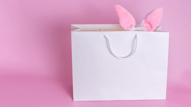 Funny ears of the Easter bunny stick out of a large white paper gift bag standing on a pink background. Copy space. Happy Easter concept