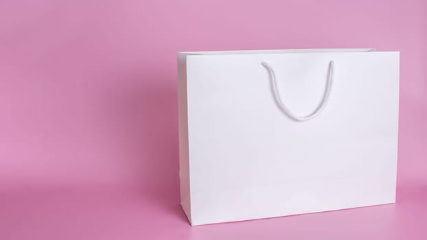 A clean large white paper bag with white rope handles stands on a pink background in close-up. Copy space. Mock up.