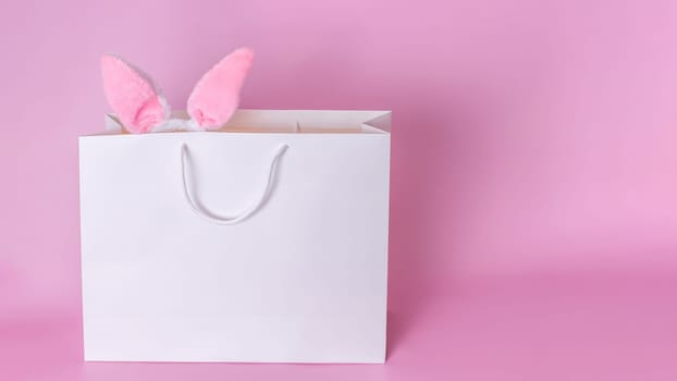 Funny pink ears of the Easter bunny stick out of a large white paper gift bag standing on a pink background. Copy space. Happy Easter concept
