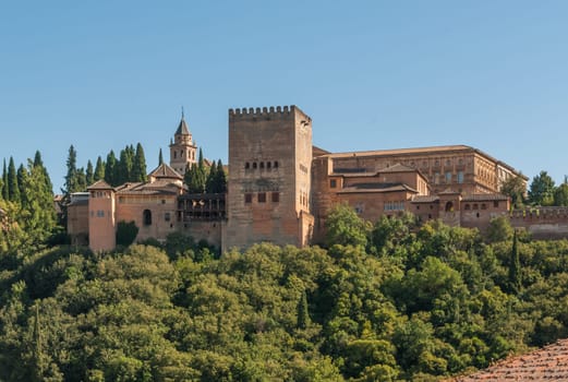 Panoramic view of the famous Alhambra in Granada/Spain, surrounded by green trees and bushes. Taken in afternoon, light blue sky in background.