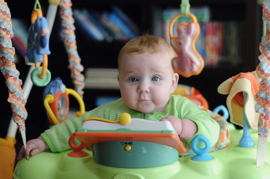 Editorial cute attentive intelligent young baby surrounded by toys in a green outfit sitting looking quizzically at the camera
