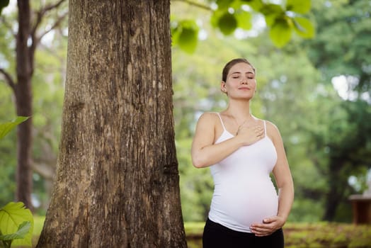 Portrait of white beautiful pregnant woman doing yoga exercise in park near tree, touching belly and heart
