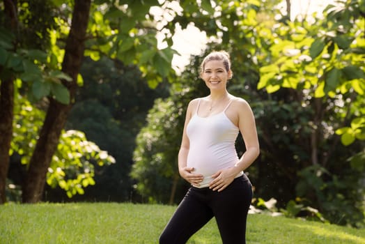 Portrait of happy pregnant woman touching belly and standing at camera smiling in city park