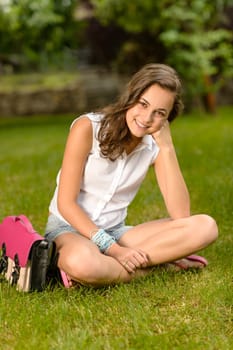 Smiling teenage girl sitting on grass with pink satchel summer