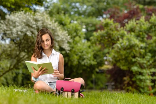 Teenage girl reading book sitting on grass summer park