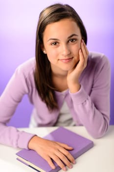 Cute student girl with book sitting behind desk purple background