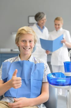 Teenager patient sitting at dental surgery thumbup dentist assistant background