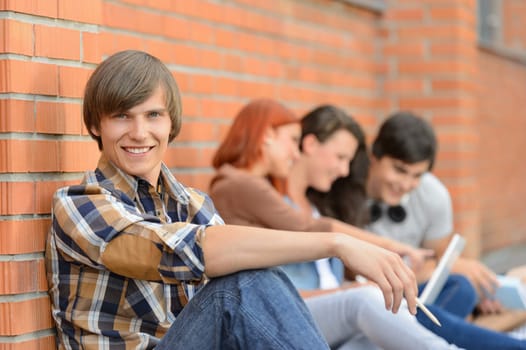 Student boy sitting by brick wall friends smiling in background