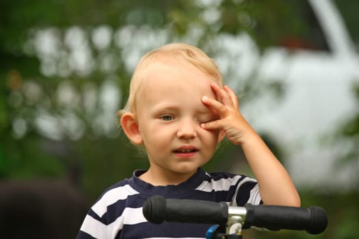 The blond boy in the striped shirt on a scooter is covering eye by hand.