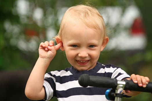 Smiling blond boy in the striped shirt on a scooter is sticking finger in ear