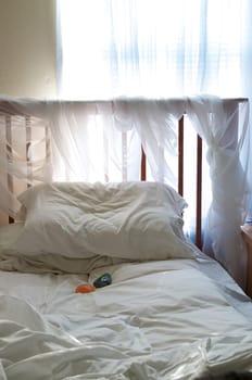 A simple bedroom under ambient light from the window coming in through blue sheer curtains to light up the sheer fabric wrapped around the headboard of the bed. There are two healing stones placed in the center of the bed.