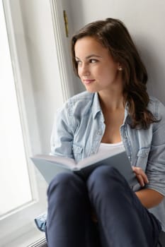 Teenage student girl sitting with book by window looking away daydreaming
