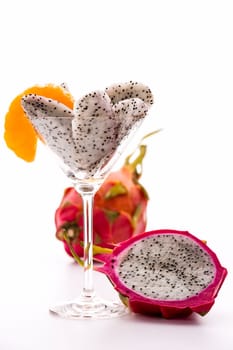 Pieces of pitaya pulp assorted in a cocktail glass and decorated with a mandarine slice. Next to it is a halved,and behind it,an entire Fruit..